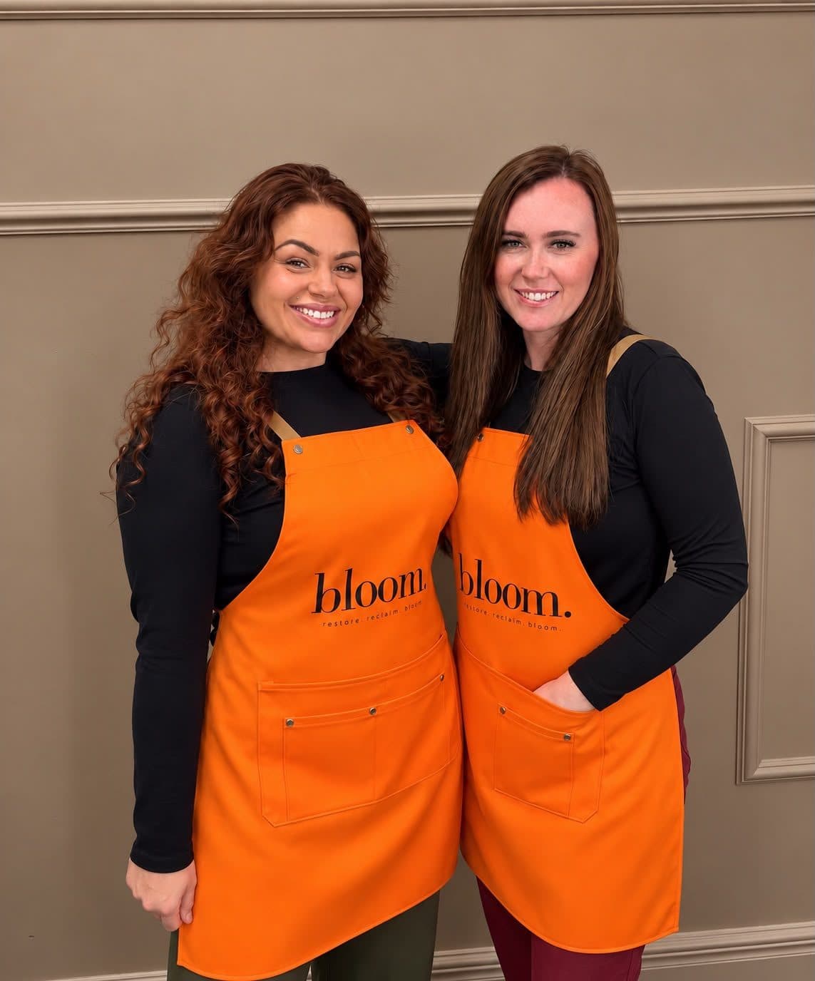 Amanda Jo and Megan Jones, Bloom's artists, smiling side by side in marigold Bloom aprons.