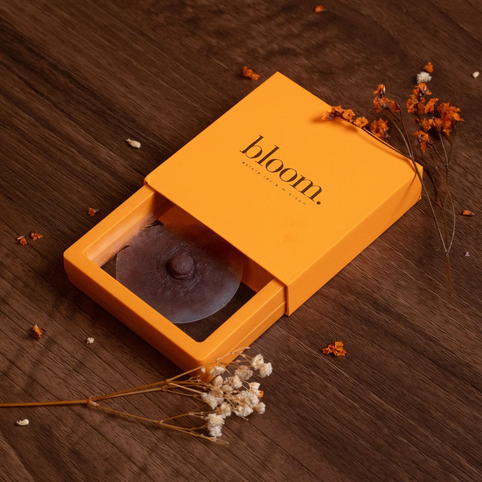 A Bloom piece in a deep shade in its open marigold box on dark wood, with dried flowers.